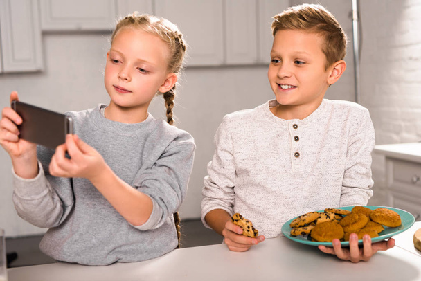 adorable child taking selfie on smartphone with brother holding plate with delicious cookies in kitchen - Foto, Bild