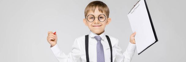 Um menino encantador com uma camisa branca, suspensórios, uma gravata e jeans leves fica em um fundo cinza. O menino segura uma caneta e lençóis para anotações
 - Foto, Imagem
