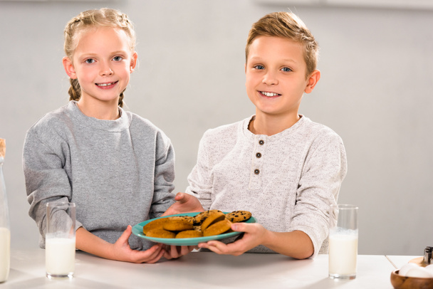 cheerful happy brother and sister holding plate with cookies near table with milk glasses  - Фото, изображение