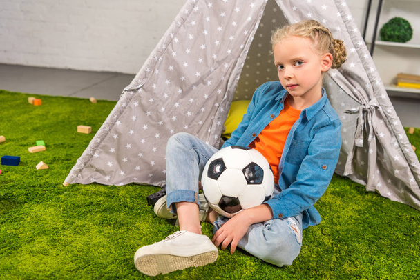 criança com bola de futebol olhando para a câmera enquanto sentado no gramado verde perto da barraca em casa
 - Foto, Imagem