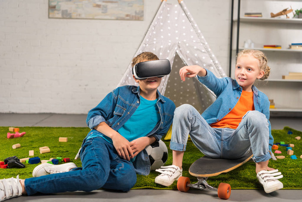 boy using virtual reality headset  and his sister pointing by finger while sitting on skateboard near wigwam at home - Fotó, kép