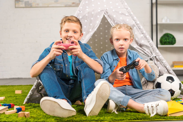 adorable sister and brother playing with joysticks near wigwam at home - Foto, afbeelding