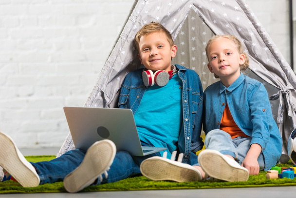 heureux petits enfants avec ordinateur portable regardant caméra et assis avec tente à la maison
 - Photo, image