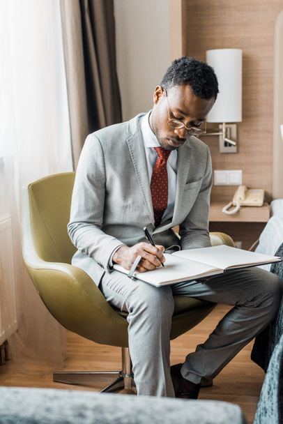 komea afrikkalainen amerikkalainen liikemies harmaassa puvussa kirjoittaa päiväkirjaan hotellihuoneessa
 - Valokuva, kuva