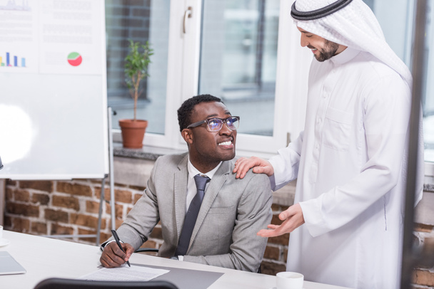 Afrikanisch-amerikanischer Geschäftsmann hält Stift in der Hand und blickt auf arabischen Partner im Amt  - Foto, Bild