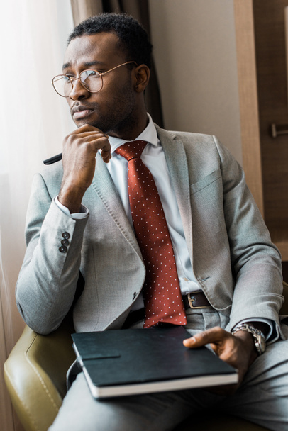 nachdenklicher afrikanisch-amerikanischer Geschäftsmann im grauen Anzug sitzt im Sessel mit Tagebuch im Hotelzimmer - Foto, Bild