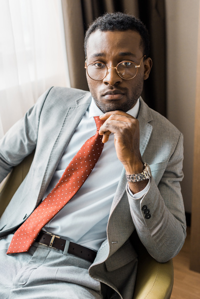 pensive african american businessman in gray suit and red tie sitting in armchair  - 写真・画像
