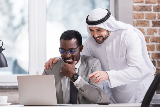 Empresarios multiétnicos mirando a la computadora portátil y sonriendo en la oficina
  - Foto, imagen