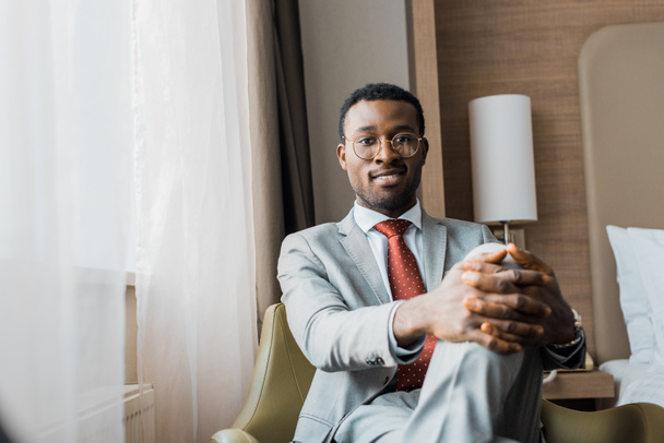 sonriente hombre de negocios afroamericano en traje gris sentado en sillón en la habitación de hotel
 - Foto, Imagen
