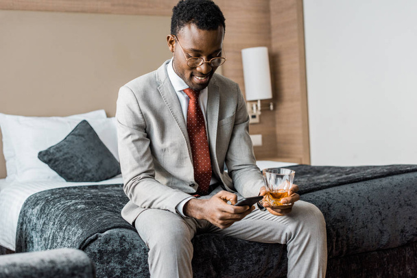усміхнений афроамериканський бізнесмен зі склянкою коньяку за допомогою смартфона, сидячи на ліжку в готелі
 - Фото, зображення