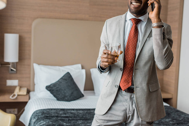 vista recortada del alegre hombre de negocios afroamericano con vaso de whisky hablando en el teléfono inteligente en el hotel
 - Foto, Imagen