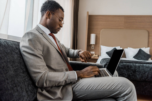 hombre de negocios afroamericano mirando reloj de pulsera mientras usa el ordenador portátil en la habitación de hotel
 - Foto, Imagen
