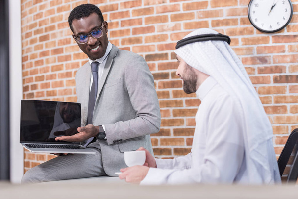 Afrikanisch-amerikanischer Geschäftsmann zeigt arabischem Geschäftspartner Laptop  - Foto, Bild