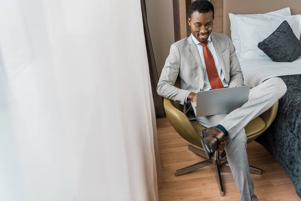hymyilevä afrikkalainen amerikkalainen liikemies puvussa työskentelee kannettavan hotellin huoneessa
 - Valokuva, kuva