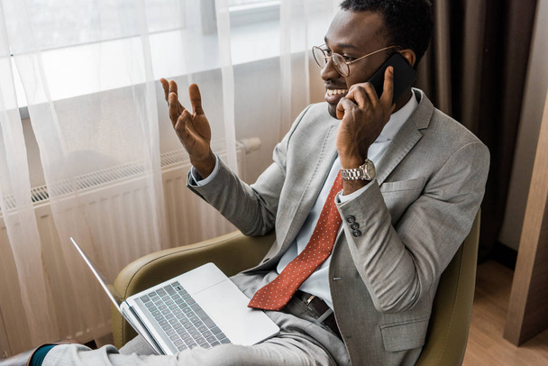 sonriente hombre de negocios afroamericano gesto, hablando en el teléfono inteligente y el uso de la computadora portátil en la habitación de hotel
 - Foto, Imagen
