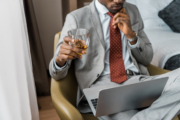 vista recortada de empresario afroamericano pensativo con vaso de whisky mirando portátil en habitación de hotel
 - Foto, Imagen