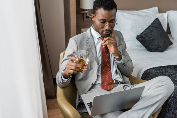 pensativo hombre de negocios afroamericano con vaso de coñac mirando portátil en habitación de hotel
 - Foto, Imagen
