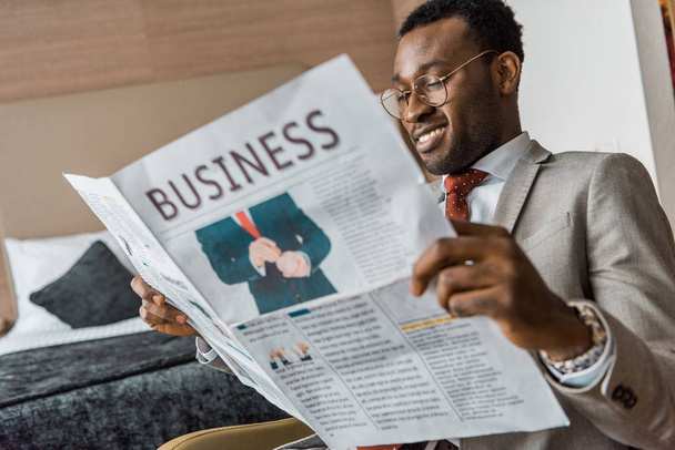 lachende Afro-Amerikaanse zakenman in pak lezen van zakelijke krant in hotelkamer - Foto, afbeelding