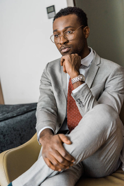 pensativo hombre de negocios afroamericano en chaqueta gris sentado en la habitación de hotel
 - Foto, Imagen