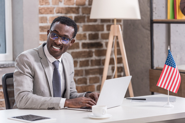 Afrikkalainen amerikkalainen liikemies käyttää kannettavaa tietokonetta ja hymyilee toimistossa
  - Valokuva, kuva