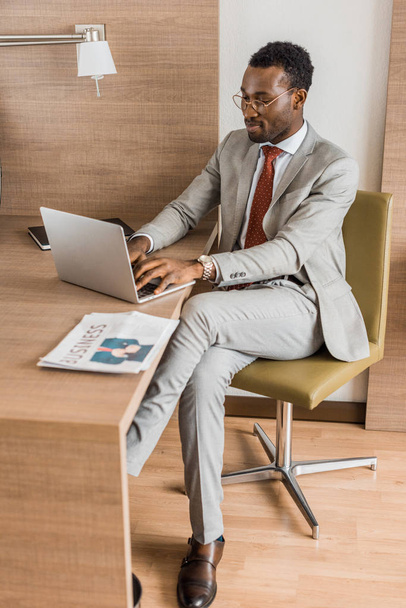 африканських американський бізнесмен в костюмі працює на ноутбуці столом з газети в готельному номері - Фото, зображення