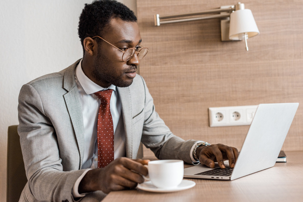Afro-Amerikaanse zakenman met koffie bezig met laptop in hotelkamer - Foto, afbeelding