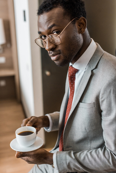guapo afroamericano hombre de negocios sosteniendo taza de café en habitación de hotel
 - Foto, Imagen