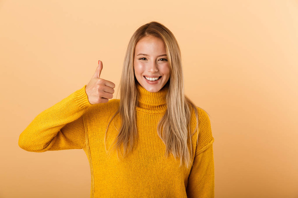 Portrait of a lovely young woman dressed in sweater standing isolated over yellow background, giving thumbs up - Foto, imagen
