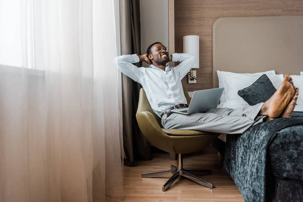smiling young african american businessman relaxing in hotel room with laptop - Zdjęcie, obraz