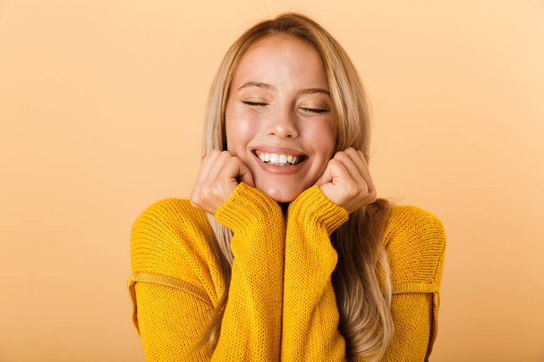Portrait of a lovely young woman dressed in sweater standing isolated over yellow background, holding hands at her face, eyes closed - Zdjęcie, obraz