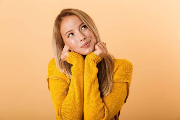 Portrait of a lovely young woman dressed in sweater standing isolated over yellow background, holding hands at her face, looking away - 写真・画像