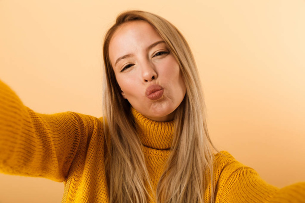 Portrait of a lovely young woman dressed in sweater standing isolated over yellow background, taking a selfie - Zdjęcie, obraz