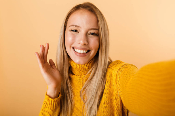 Πορτρέτο του ένα αστείο νεαρή γυναίκα ντυμένη με πουλόβερ, στέκεται απομονωθεί σε κίτρινο φόντο, λαμβάνοντας μια selfie - Φωτογραφία, εικόνα