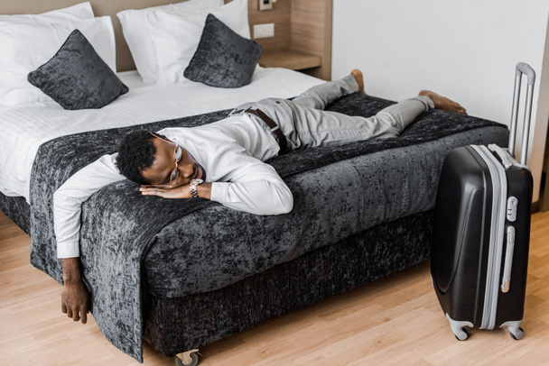 väsynyt afrikkalainen amerikkalainen mies muodollinen kulumista nukkuu sängyllä hotellihuoneessa matkalaukku
 - Valokuva, kuva