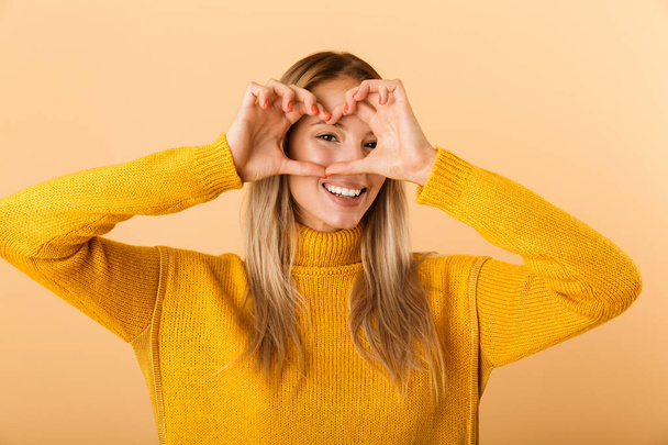 Portrait of a happy young woman dressed in sweater standing isolated over yellow background, showing love gesture - Valokuva, kuva