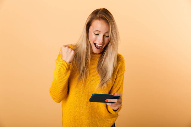 Portrait of a happy young woman dressed in sweater standing isolated over yellow background, playing games on mobile phone, celebrating - Fotografie, Obrázek