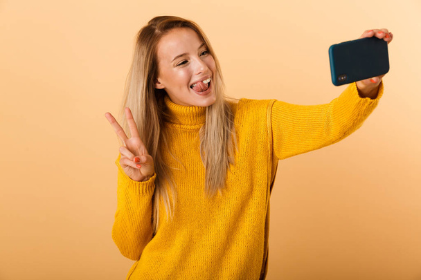 Portrait of a funny young woman dressed in sweater standing isolated over yellow background, taking a selfie, showing peace - Фото, изображение