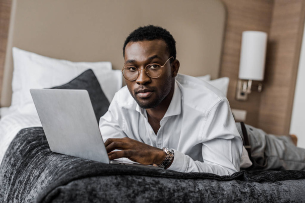 komea afrikkalainen amerikkalainen liikemies silmälaseissa työskentelee kannettavan tietokoneen kanssa hotellivuoteella
 - Valokuva, kuva
