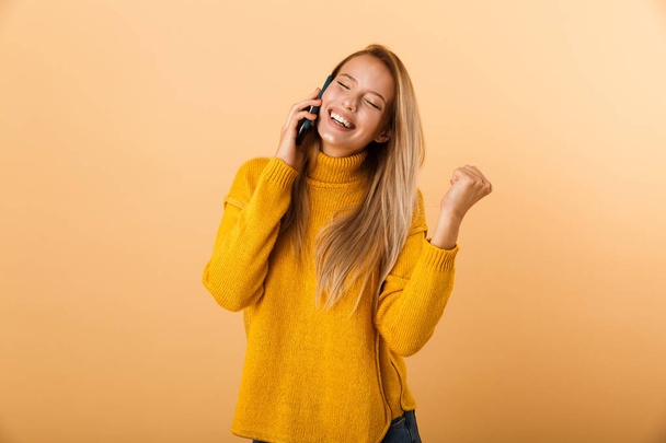 Portret podekscytowany młoda kobieta ubrana w sweter stojąc na białym tle nad żółtym tle, rozmowy na telefon komórkowy, świętować sukces - Zdjęcie, obraz