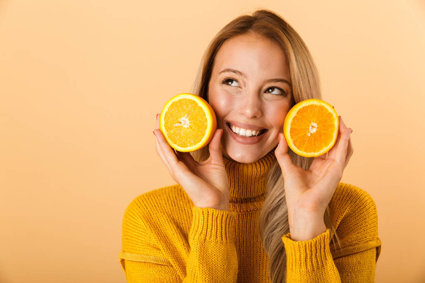 Portrait of a smiling young woman dressed in sweater standing isolated over yellow background, holding sliced orange at her face - Valokuva, kuva