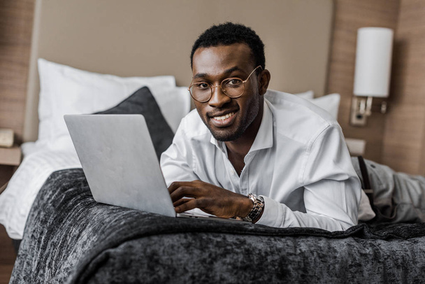 smiling african american businessman in eyeglasses working with laptop on hotel bed - Fotoğraf, Görsel