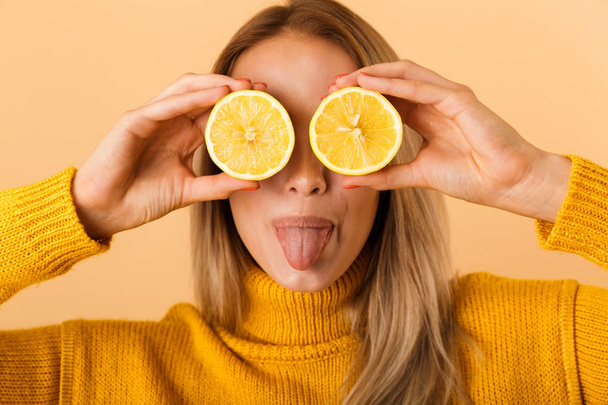 Photo of a beautiful woman covering eyes with citrus lemons posing isolated over yellow wall background. - Foto, immagini