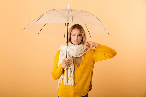 Photo of a displeased woman wearing scarf holding umbrella posing isolated over yellow wall background showing thumbs down gesture. - Valokuva, kuva