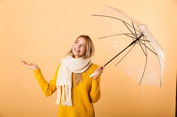 Photo of a cute woman wearing scarf holding umbrella posing isolated over yellow wall background. - Fotó, kép