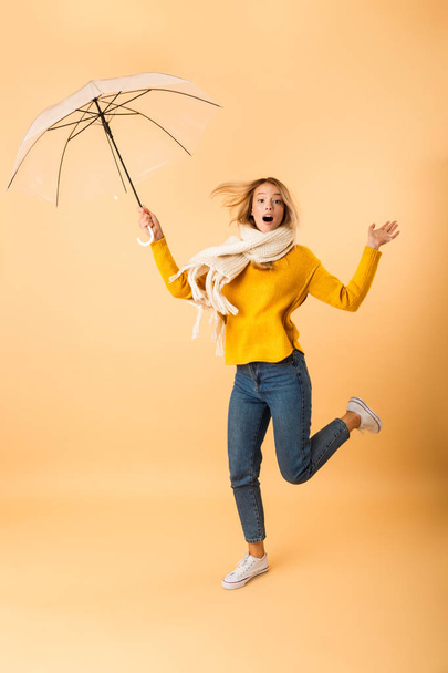Foto de una linda mujer con bufanda sosteniendo paraguas saltando aislada sobre fondo amarillo de la pared
. - Foto, Imagen
