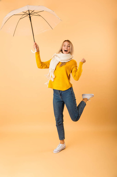 Photo of a cute woman wearing scarf holding umbrella jumping isolated over yellow wall background. - 写真・画像