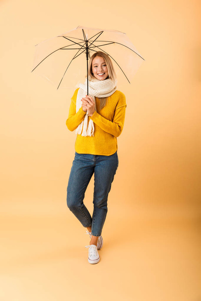 Photo of a cute woman wearing scarf holding umbrella posing isolated over yellow wall background. - 写真・画像