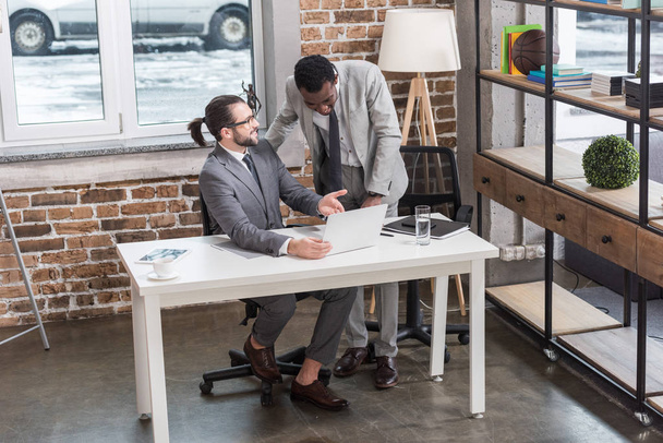 przystojny wieloetnicznym biznesmenów, uśmiechając się i patrząc na laptopie w nowoczesnym biurze - Zdjęcie, obraz