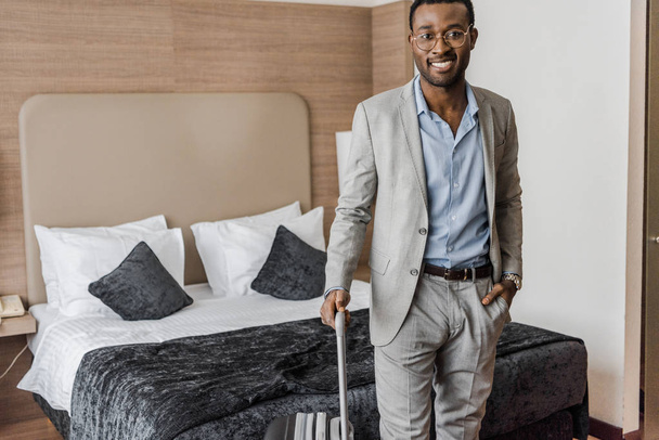 handsome smiling african american businessman with suitcase in hotel room - Foto, imagen