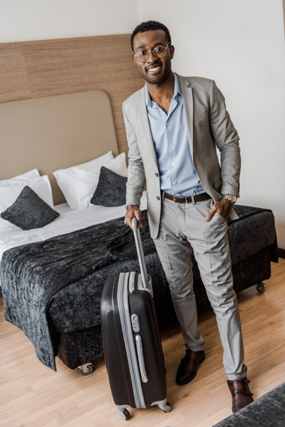 ベッドとホテルの部屋でスーツケースにスーツでアフリカ系アメリカ人のビジネスマン - 写真・画像